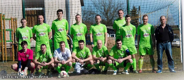 Eine Gruppe von Männer in grünen T-Shirts steht auf einem Fußballfeld, einige hocken, mit einem Ball vor ihnen und einem Torpfosten rechts, vor einer Kulisse aus Bäumen, Gebäuden und Fahrzeugen.