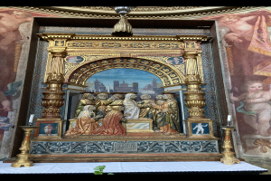 Ein Altar in einer Kirche mit Skulpturen, Kerzen und anderen Gegenständen, vor einer Wand mit einem Bild, und einer Pflanze unten.