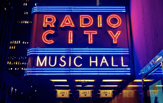 Außenansicht des Radio City Music Hall in New York City bei Nacht, hell erleuchtet und von hohen Gebäuden umgeben, mit einem "Radio City"-Schild und zusätzlichen Schildern auf der rechten Seite.