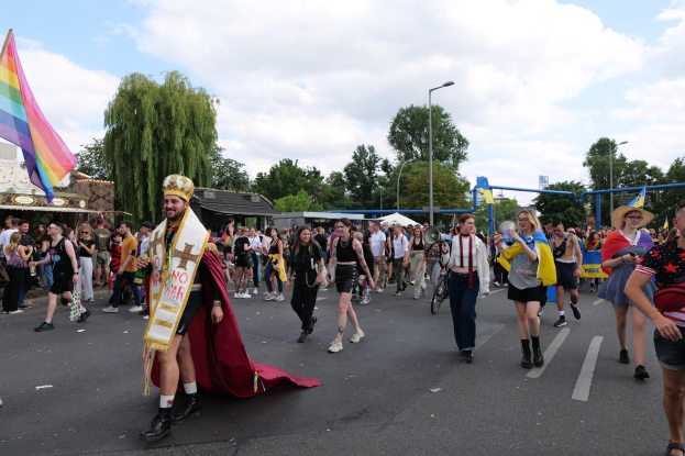 Menschen marschieren in einem Umzug, einige halten Musikinstrumente und andere tragen Mützen, tragen eine Regenbogenfahne, mit Laternenmasten, Bäumen, Hütten und einem bewölkten Himmel im Hintergrund.