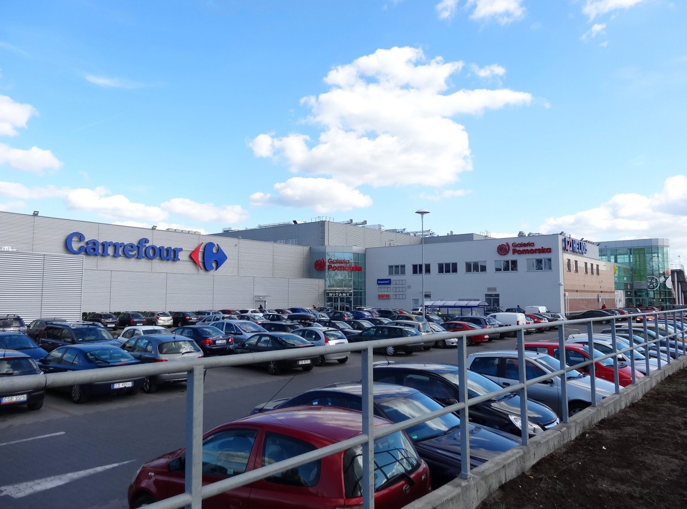 Großer Autohandel mit mehreren parkenden Fahrzeugen vor einem städtischen Hintergrund mit Gebäuden, Straßenlaternen, Schildern, fahrenden Autos und einer bewölkten Himmel.