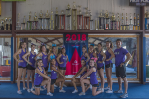Gruppe von Cheerleadern in lila T-Shirts und schwarzen Shorts, die mit einer Vitrine voller Trophäen posieren, einige stehen und einige hocken, mit einem "2018 Summit Cheerleading Champions"-Banner im Hintergrund.