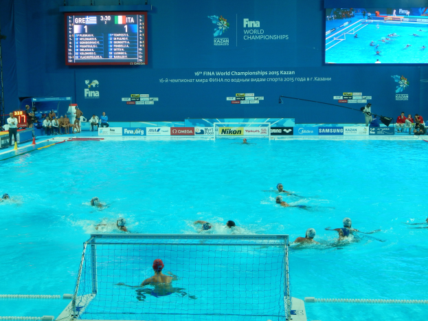 Gruppe von Athleten beim Wasserballspielen in einem Pool während der FINA-Weltmeisterschaften 2016 in Kazan, mit Zuschauern und Anzeigetafel im Hintergrund.