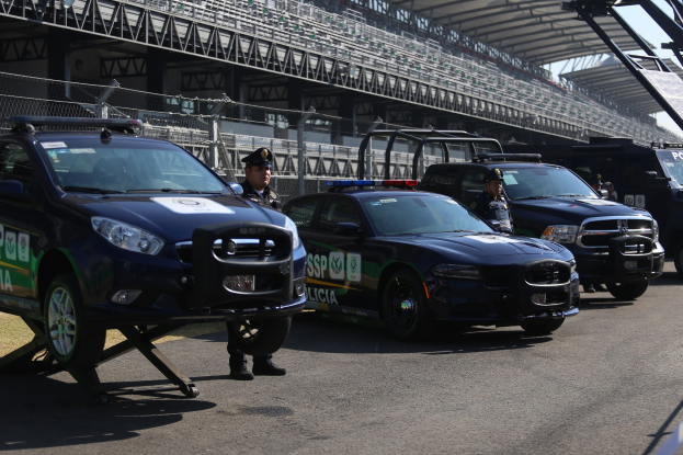 Polizeiautos vor einer Rennstrecke geparkt, uniformierte Personen, Stadion im Hintergrund und Maschendraht unter einem sichtbaren Himmel.