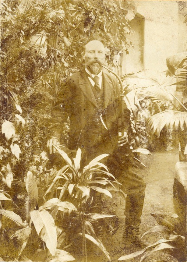 Altes Foto eines Mannes in Anzug und Krawatte, der in einem Garten mit Pflanzen, Bäumen und einer Wand im Hintergrund steht.