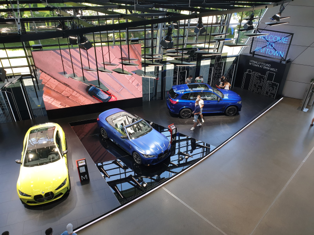 Eine Gruppe von Menschen im BMW Museum in München, Deutschland, mit Autos auf der linken Seite und einem Schild mit der Aufschrift "BMW Museum München" auf der rechten Seite, das auf eine städtische Außenansicht durch Glasfenster blickt.