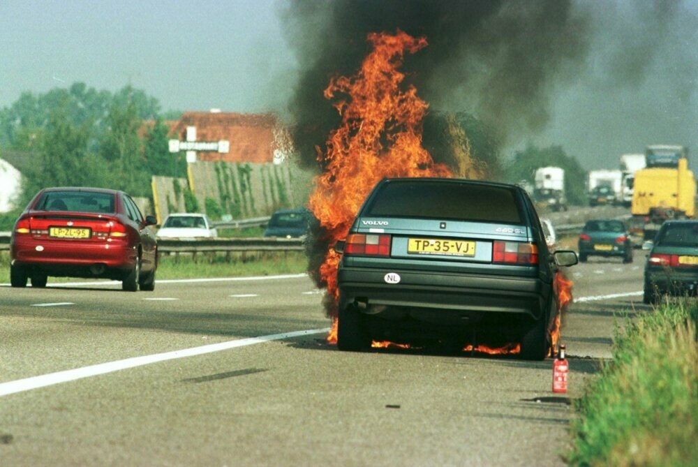 Ein Auto in Flammen auf der Seite der Stra├če, mit umgebenden Fahrzeugen, B├Ąumen, Geb├Ąuden, einem klaren blauen Himmel, Gras und einem Feuerl├Âscher.