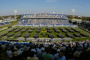 Eine große Menschenmenge in einem Stadion bei einem Fußballspiel, wobei einige stehen und andere sitzen, auf einem grünen Feld unter einem klaren blauen Himmel, umgeben von Laternenmasten, Bäumen und Gebäuden.