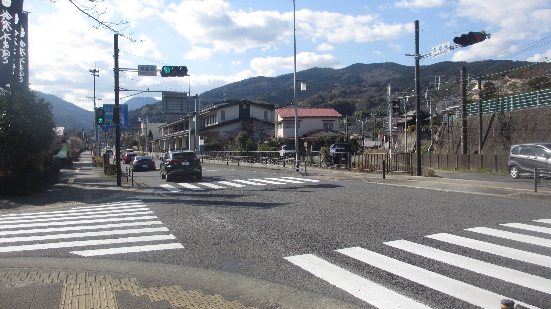 Stadtstraße mit fahrenden Autos, Zebrastreifen, Strommasten mit Drähten, Ampeln, Gebäude mit Fenstern, Bäume, entfernte Hügel und ein bewölkter Himmel.