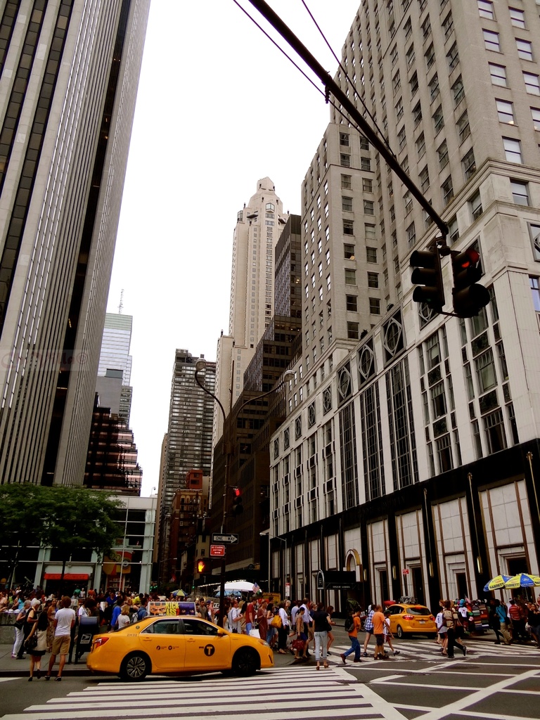 Eine Stadtansicht mit Gebäuden, Ampeln, Fenstern, wenigen Passanten, Autos und Himmel.