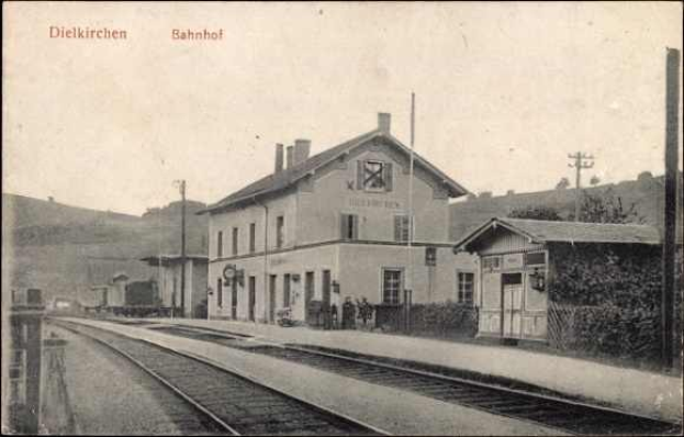 Schwarz-weiß-Fotografie eines Bahnhofs in Dielkirchen, Deutschland, mit einem Zug auf den Gleisen, umgeben von Häusern, Strommasten, Bäumen, Pflanzen, Menschen, fernen Hügeln und einem klaren Himmel, mit Stationsnamen-Text oben und unten.