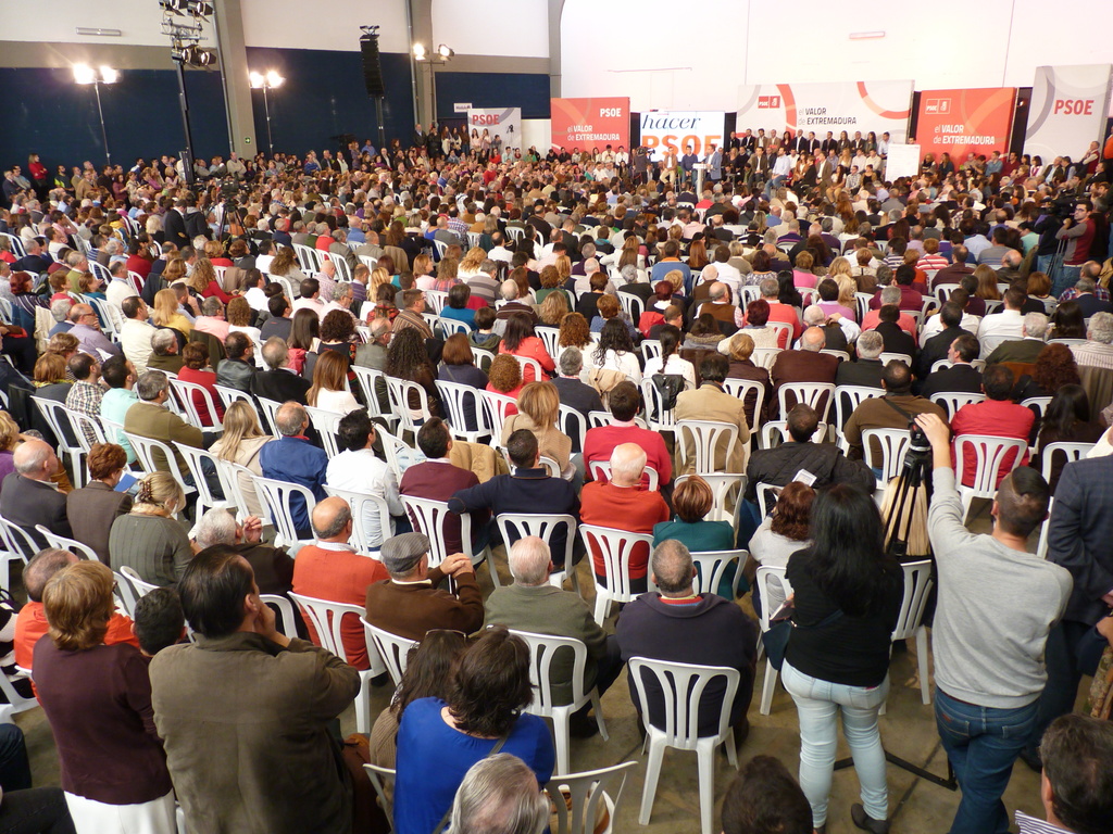 Ein volles Stadion mit vielen sitzenden und stehenden Menschen, eine Bühne mit zwei Rednern am Podium und verschiedene Banner und Lichter in der Nähe.