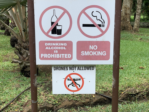 Schild am Straßenrand mit der Aufschrift "Alkoholtrinken ist verboten" und "Rauchen verboten", umgeben von Bäumen und Gras.