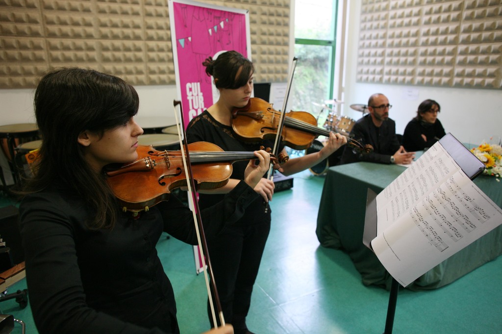 Zwei Frauen, die Geige spielen, in einem Raum mit einem Musikständer, einem Tisch, zwei sitzenden Personen und einer Tafel im Hintergrund.