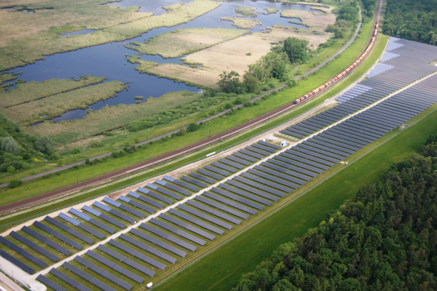 Luftaufnahme einer Solar-Farm mit Panels, einem Zug auf einer Schiene, umgeben von Bäumen, Gras, Wasser und Vegetation.