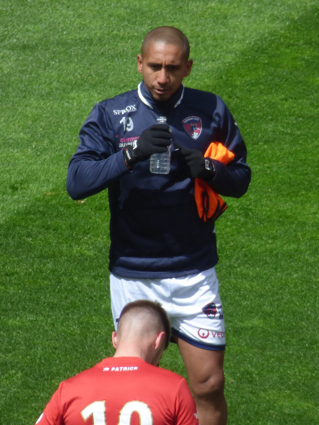 Ein Mann in einem Sportdress steht auf einem grünen Feld und hält in einer Hand eine Flasche und in der anderen ein oranges Tuch.