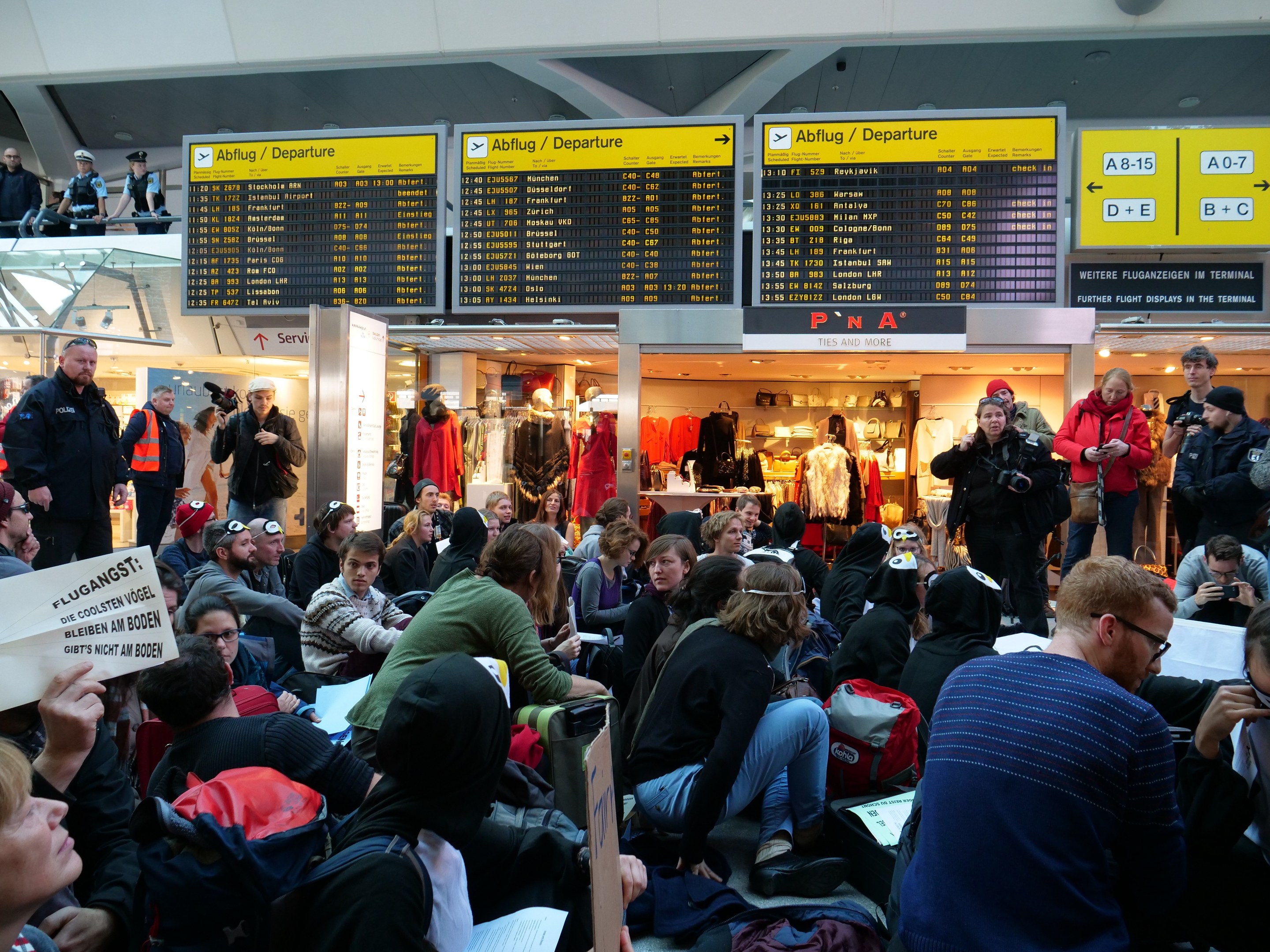 Menschen sitzen und stehen in einem Flughafen während einer Demonstration, mit Informationsschildern, Schaufensterpuppen und Deckenleuchten im Hintergrund.