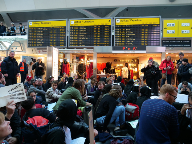 Menschen sitzen und stehen in einem Flughafen während einer Demonstration, mit Informationsschildern, Schaufensterpuppen und Deckenleuchten im Hintergrund.