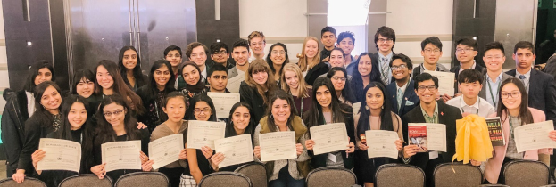 Gruppe von Menschen mit Urkunden in einem Raum mit Stühlen und einer Wand im Hintergrund, wahrscheinlich feiern sie eine Abschlussfeier für das Studienjahr 2019-2020.