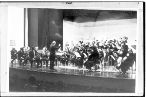 Ein Schwarz-Weiß-Foto eines Dirigenten in Anzug und Krawatte, der auf einer Bühne steht und ein Orchester mit verschiedenen Instrumenten wie Geigen, Celli und Flöten leitet, während das Publikum auf Stühlen aufmerksam zuhört.