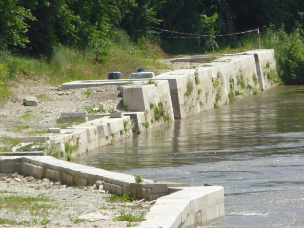 Ein ruhiger, klarer Fluss mit einer zentralen Rückhaltewand, umgeben von üppiger Vegetation und Bäumen, die den Himmel widerspiegeln.