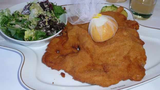 Ein weißer Teller mit einem goldbraunen Schnitzel, einer Salatschüssel und einem Glas Getränk auf einem mit einer Decke bedeckten Tisch.
