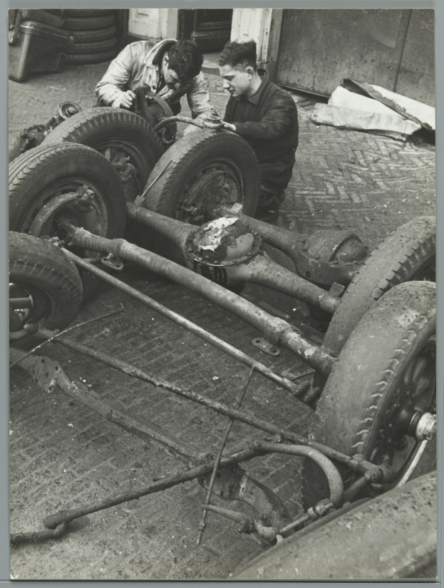 Zwei Männer in einer Garage, die Werkzeuge halten, arbeiten an einem Auto umgeben von verstreuten Reifen und Gegenständen, dargestellt in Schwarz-Weiß.