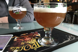 Zwei Gläser goldfarbenes Bier auf einem Tisch mit einem Handy in der Nähe, umgeben von Menschen und Rahmen an der Wand im Hintergrund.