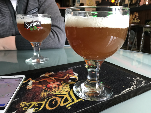 Zwei Gläser goldfarbenes Bier auf einem Tisch mit einem Handy in der Nähe, umgeben von Menschen und Rahmen an der Wand im Hintergrund.