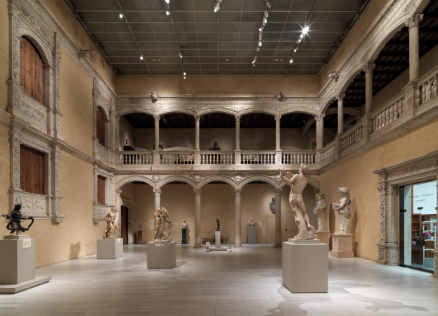 Großer Raum in einem Museum mit zahlreichen Statuen und Skulpturen auf Sockeln, architektonischen Elementen wie Wänden, Säulen und Geländern, einer beleuchteten Decke und einigen Besuchern.