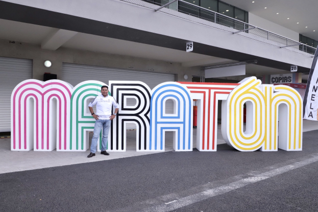Ein Mann steht am Start eines Marathons vor einem großen "Marathon"-Schild mit einem Banner rechts daneben, im Hintergrund ein Gebäude mit Lampen, Geländern und Fensterläden.