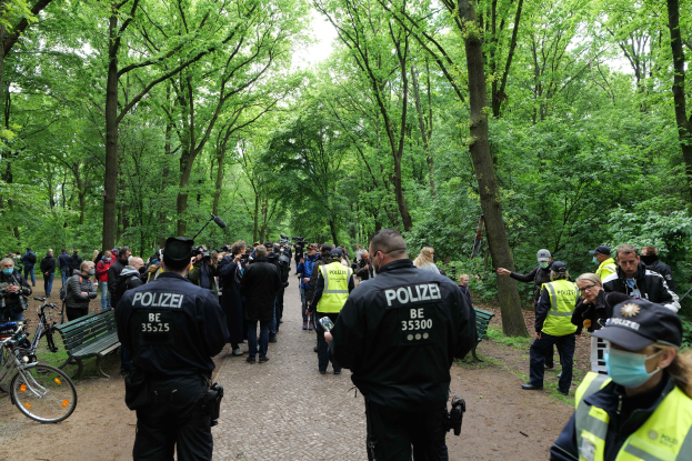 Polizeibeamte in Uniform vor einer Menschenmenge mit Fahrrädern und einer Bank im Vordergrund und Bäumen im Hintergrund.
