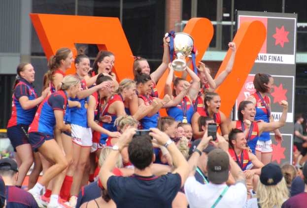 Gruppe von Frauen-AFL-Spielerinnen feiert mit einem Pokal, einige halten ihn in die Höhe, während andere Fotos machen, vor einer jubelnden Menge mit Anzeigetafeln und einem Glasgebäude im Hintergrund.