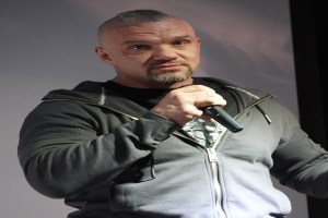 Ein Mann in einer schwarzen Kapuzenjacke, einem schwarzen Anzug und einem weißen Hemd hält ein Mikrofon und steht selbstbewusst vor einer Wand.