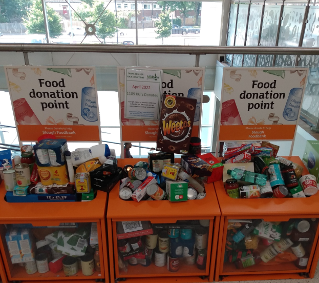 Orange Schübe mit Lebensmittelspenden, darunter Flaschen, Dosen und Kisten, mit informativen Schildern im Hintergrund; Hintergrund zeigt Fahrzeuge, Bäume, Gebäude und Himmel durch Glasfenster.