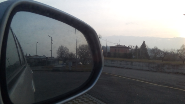 Der Seitenspiegel eines Autos am Straßenrand, der ein Gebäude, Bäume, Pfähle, Drähte und den Himmel reflektiert.