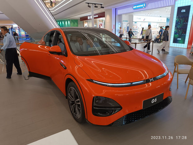Oranges Elektroauto in einer Ausstellungshalle umgeben von einer Gruppe von Menschen, mit Stühlen und einem Wagen rechts und Geschäften im Hintergrund.