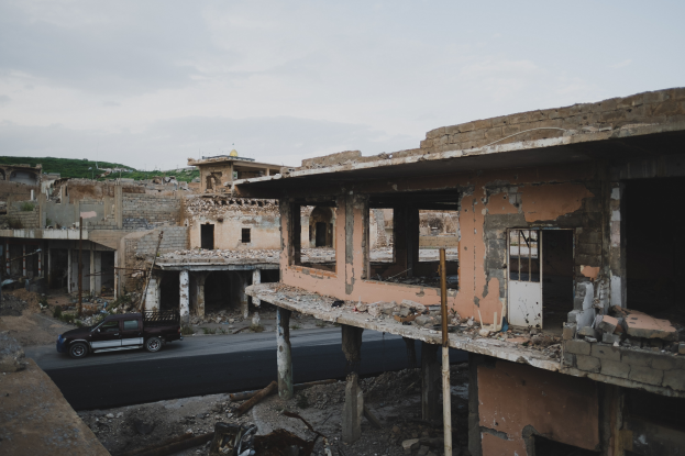 Ein Lastwagen fährt eine Straße neben einem zerstörten Gebäude mit gebrochenen Wänden und verstreuter Trümmer, mit anderen Gebäuden, Polen, Bäumen, einem Hügel und dem Himmel im Hintergrund.