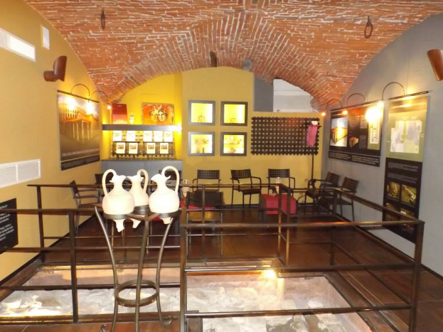 Ein Museumsraum voller Tische, Stühle, Geländer, Vasen, Texttafeln, Wandrahmen, Lampen und diverser Ausstellungsstücke.